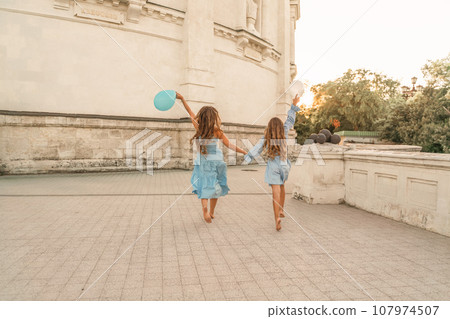 Daughter mother run holding hands. In blue dresses with flowing long hair, they hold balloons in their hands against the backdrop of a sunset and a white building. 107974507