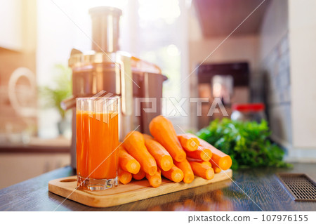Glass of freshly squeezed carrot juice, carrots and juicer in the kitchen Glass of freshly squeezed carrot juice, carrots and juicer in the kitchen 107976155