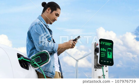 Asian man using smartphone while electric car recharging energy. Peruse Asian man using smartphone while electric car recharging energy. Peruse 107976759