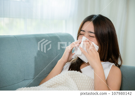People sick. Ill Asian young woman cold covered with blanket lying on sofa and sneeze with tissue paper at home, Female sick allergic blowing nose sneezing in tissue at home, Flu health care 107980142
