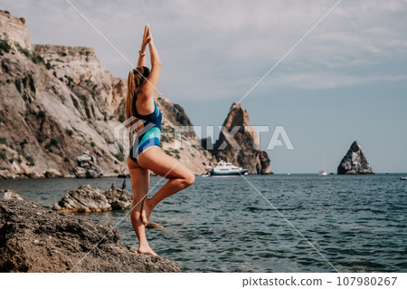 Woman sea yoga. Back view of free calm happy satisfied woman with long hair standing on top rock with yoga position against of sky by the sea. Healthy lifestyle outdoors in nature, fitness concept Woman sea yoga. Back view of free calm happy satisfied woman with long hair standing on top rock with yoga position against of sky by the sea. Healthy lifestyle outdoors in nature, fitness concept 107980267