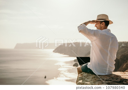 Digital nomad, man in the hat, a businessman with a laptop sits on the rocks by the sea during sunset, makes a business transaction online from a distance. Remote work on vacation. Digital nomad, man in the hat, a businessman with a laptop sits on the rocks by the sea during sunset, makes a business transaction online from a distance. Remote work on vacation. 107980340