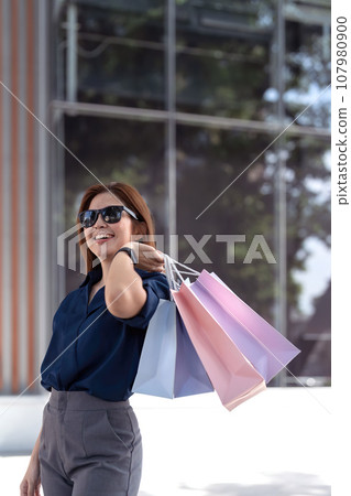 Happy beautiful young stylish woman with shopping bags while walking near mall on holiday Happy beautiful young stylish woman with shopping bags while walking near mall on holiday 107980900