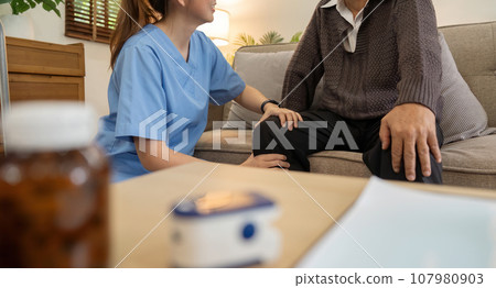 Senior elderly male or patient sitting on sofa in living room the caregiver is checking his body in home Senior elderly male or patient sitting on sofa in living room the caregiver is checking his body in home 107980903