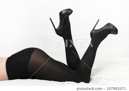 Crop view of female in black pantyhose and high heel shoes lying on front with her legs bent in knees and raised up with heels. Unrecognizable woman on bed on fleecy blanket on white background 107981071
