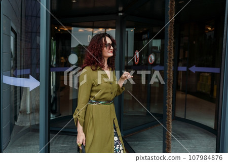 Woman leaves a supermarket. Caucasian model with long brunette hair, wears sunglasses and a khaki dress. 107984876