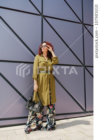 Portrait of a woman standing near a supermarket building. Caucasian model with long brunette hair, wears sunglasses and a khaki dress. Portrait of a woman standing near a supermarket building. Caucasian model with long brunette hair, wears sunglasses and a khaki dress. 107985010