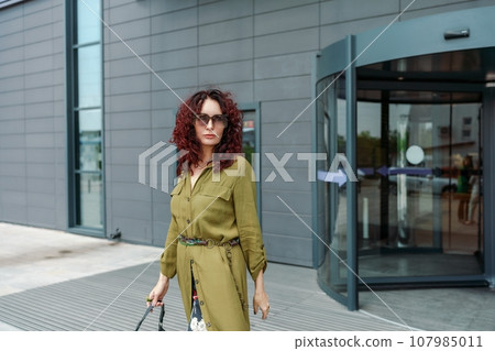 Portrait of a woman standing near a supermarket building. Caucasian model with long brunette hair, wears sunglasses and a khaki dress. Portrait of a woman standing near a supermarket building. Caucasian model with long brunette hair, wears sunglasses and a khaki dress. 107985011