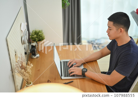 Side view of concentrated asian male freelancer working online or browsing web on laptop at home Side view of concentrated asian male freelancer working online or browsing web on laptop at home 107988555