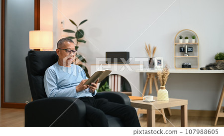 Calm senior man in casual clothes resting on armchair and reading favorite literature Calm senior man in casual clothes resting on armchair and reading favorite literature 107988605