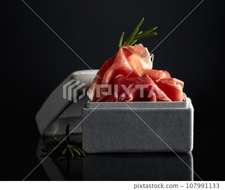 Prosciutto with rosemary in a gray gift box on a black background. Prosciutto with rosemary in a gray gift box on a black background. 107991133