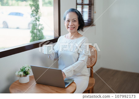 Portrait of an elderly Asian woman in modern pose doing computer work and drinking coffee 107992173