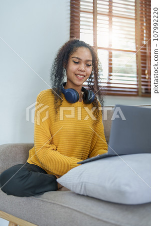Portrait of an African American sitting on the sofa wearing on-ear headphones and using a computer at home. Portrait of an African American sitting on the sofa wearing on-ear headphones and using a computer at home. 107992220