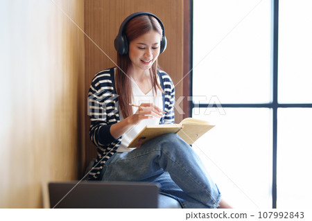 Portrait of a young Asian teenage girl in love wearing headphones, using a notebook and computer to study online via video conferencing. Portrait of a young Asian teenage girl in love wearing headphones, using a notebook and computer to study online via video conferencing. 107992843