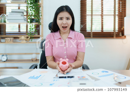 Portrait of an Asian female businesswoman showing a serious and anxious expression in an SME business , but no money in the pink pig piggy bank , saving plans. 107993149