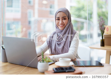 A beautiful Muslim woman with a smiling face in the morning drinking coffee and using a computer. 107993357