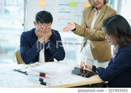 An image of an Asian male employee looking worried and sad about being scolded by his boss for failing to meet sales targets, concept of disappointment and failure in his career. 107993405