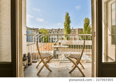 a balcony with two chairs and a table in the middle, looking out into the yard from inside an open door 107993905