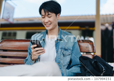 Asian teenage male traveler using her smart phone mobile while waiting for a train at a station 107994151