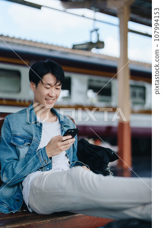 Asian teenage male traveler using her smart phone mobile while waiting for a train at a station Asian teenage male traveler using her smart phone mobile while waiting for a train at a station 107994153