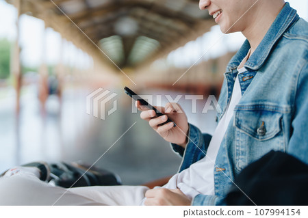 Asian teenage male traveler using her smart phone mobile while waiting for a train at a station Asian teenage male traveler using her smart phone mobile while waiting for a train at a station 107994154