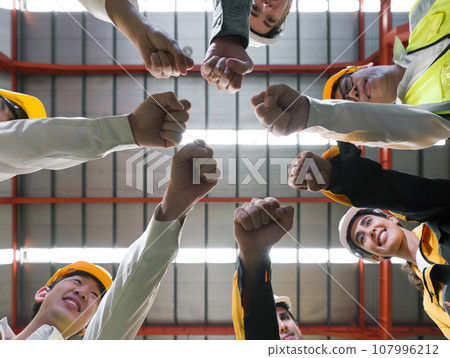 Group of male and female factory labor bump their fists together after finish meeting. Everyone wearing safety uniform and helmet. Workers working in the metal sheet factory. Bottom View 107996212