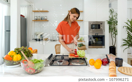 Young housewife slice bell pepper on a wooden chop board. The counter displays vibrant, fresh produce, revealing the beginnings of a nutritional meal. Young housewife slice bell pepper on a wooden chop board. The counter displays vibrant, fresh produce, revealing the beginnings of a nutritional meal. 107996271