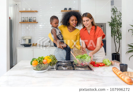 Two young diverse lesbian women spending time in a kitchen at home, harmoniously sharing food preparation duties whilst simultaneously nurturing a baby. Two young diverse lesbian women spending time in a kitchen at home, harmoniously sharing food preparation duties whilst simultaneously nurturing a baby. 107996272