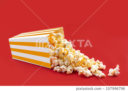 Tasty cheese popcorn falling out of a yellow striped carton bucket, isolated on red background 107996796