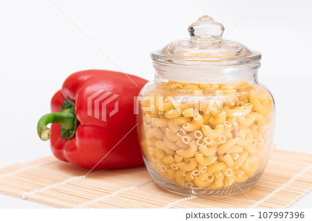 Uncooked Chifferi Rigati Pasta in Glass Jar and Red Bell Pepper on Bamboo Mat 107997396