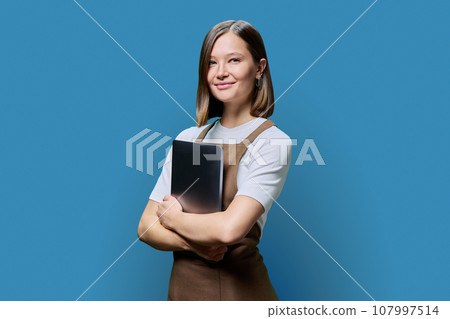 Portrait of young woman in apron holding laptop on blue background Portrait of young woman in apron holding laptop on blue background 107997514