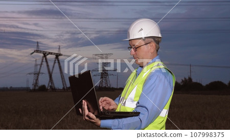 Mature electrician with laptop inspects network of powerlines in evening field. Engineer conducts diagnostics of power generation substation facilities with laptop in sunset country field Mature electrician with laptop inspects network of powerlines in evening field. Engineer conducts diagnostics of power generation substation facilities with laptop in sunset country field 107998375