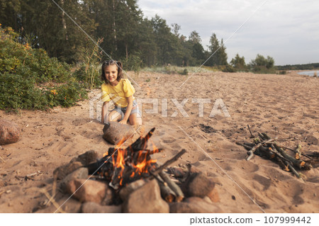 happy smiling cheerful girl 6 years old sits sitting on beach near forest opposite fire and enjoys in summer day, picnic, vacation happy smiling cheerful girl 6 years old sits sitting on beach near forest opposite fire and enjoys in summer day, picnic, vacation 107999442