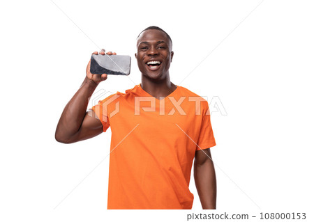 young authentic african man in orange t-shirt rejoices showing smartphone screen with mockup 108000153