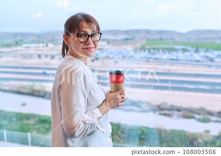 Woman looking out airport window with glass of takeaway coffee 108003508