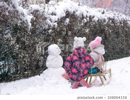 Two sisters on sled looking to snowman at snow day in park. Two sisters on sled looking to snowman at snow day in park. 108004158