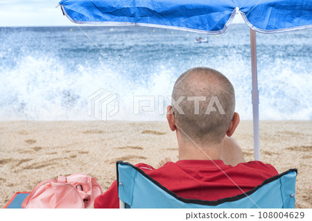 Unknown Man sits on a sun lounger under an umbrella and looks at the sea 108004629