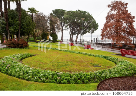 Red benches in park in centre of city Locarno with pine trees and orange (autumn leaf color) larch tree on lake Red benches in park in centre of city Locarno with pine trees and orange (autumn leaf color) larch tree on lake 108004735