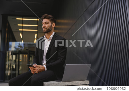 Handsome male manager in suit with phone sitting on bench with laptop near office building Handsome male manager in suit with phone sitting on bench with laptop near office building 108004982