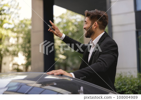 Cheerful businessman standing near car with mobile phone and waving hi to colleagues near office Cheerful businessman standing near car with mobile phone and waving hi to colleagues near office 108004989