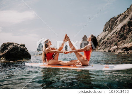 Woman sup yoga. Two happy sporty women practising yoga pilates on paddle sup surfboard. Female stretching doing workout on sea water. Modern individual female hipster outdoor summer sport activity. Woman sup yoga. Two happy sporty women practising yoga pilates on paddle sup surfboard. Female stretching doing workout on sea water. Modern individual female hipster outdoor summer sport activity. 108013453