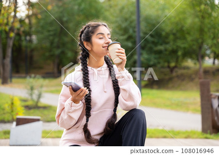 Beautiful Indian woman enjoying coffee takeaway with closed eyes while resting in autumn park, holding smartphone Beautiful Indian woman enjoying coffee takeaway with closed eyes while resting in autumn park, holding smartphone 108016946