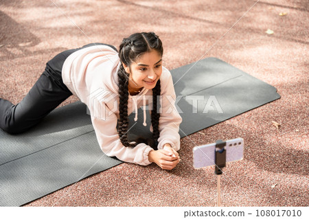 Online workout in modern outdoors sportground performed by young Indian woman doing twine and looking at smartphone 108017010