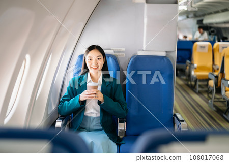 Portrait of  successful Asian businesswoman or entrepreneur in a formal suit on an airplane sitting in business class using a phone, computer laptop. Travel in style, work with grace. 108017068