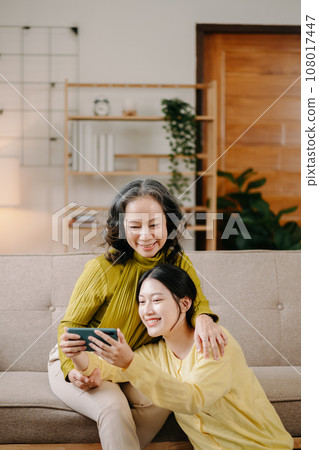 Happy adult granddaughter and senior grandmother having fun enjoying talk sit on sofa in living room at home. Happy adult granddaughter and senior grandmother having fun enjoying talk sit on sofa in living room at home. 108017447