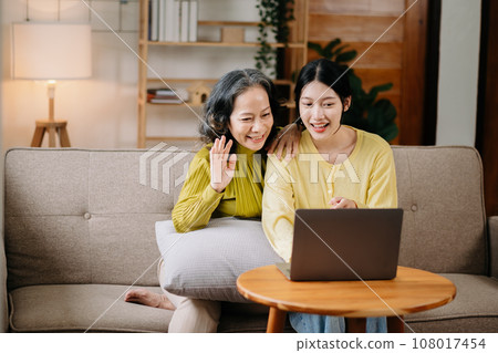 Happy adult granddaughter and senior grandmother having fun enjoying talk sit on sofa in modern living room, using a laptop, tablet bonding chatting relaxing at home together. 108017454