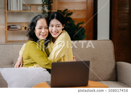 Happy adult granddaughter and senior grandmother having fun enjoying talk sit on sofa in modern living room, using a laptop, tablet bonding chatting relaxing at home together. 108017458