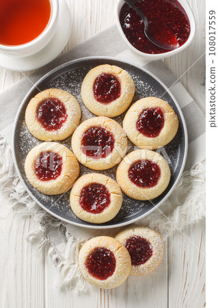 Thumbprint Christmas cookies filled with raspberry jam closeup on the plate served with tea on the wooden table. Vertical top view 108017559