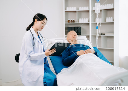 Asian doctor in white suit take notes while discussing and Asian elderly, man patient who lying on bed with receiving saline solution in hospital . Asian doctor in white suit take notes while discussing and Asian elderly, man patient who lying on bed with receiving saline solution in hospital . 108017814