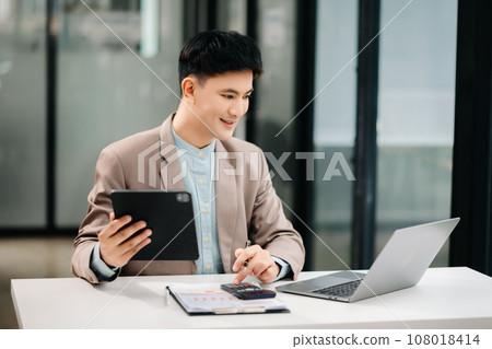 Young handsome man typing on tablet and laptop while sitting at the working wooden table modern office room.. Young handsome man typing on tablet and laptop while sitting at the working wooden table modern office room.. 108018414